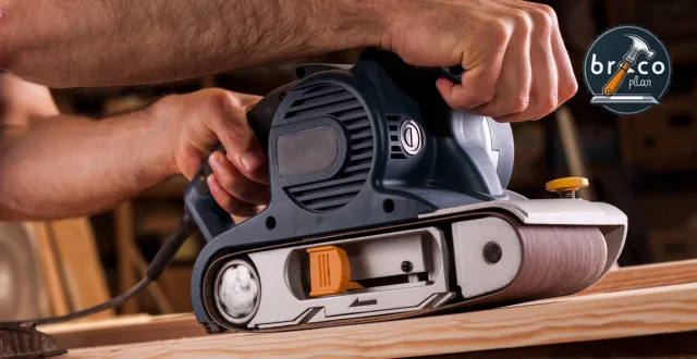 Hombre usando una lijadora de banda eléctrica sobre un tablero de madera en un taller, lijando la superficie con precisión.